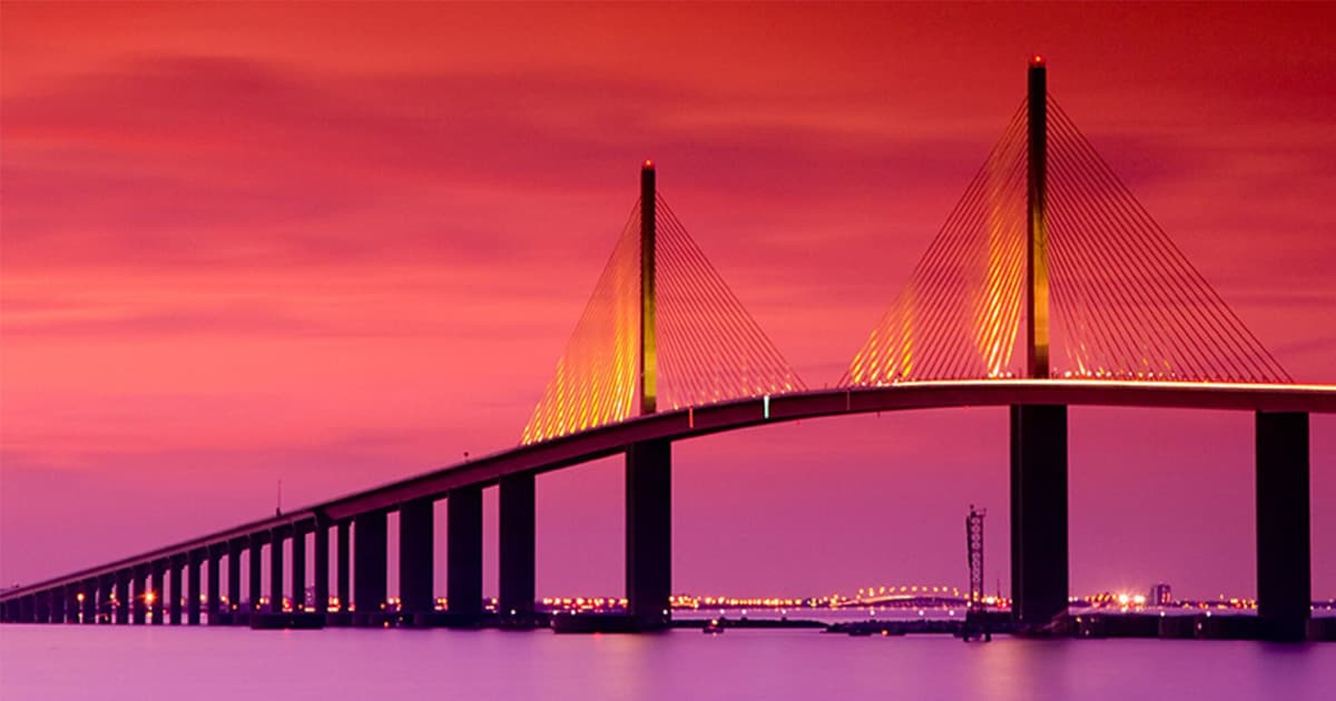 Wide profile photograph of the Sunshine Skyway Bridge crossing Tampa Bay at twilight, with the cable-stayed yellow main span illuminated against a vivid pink and magenta sunset sky and the lights of the Pinellas County shoreline reflecting on calm water